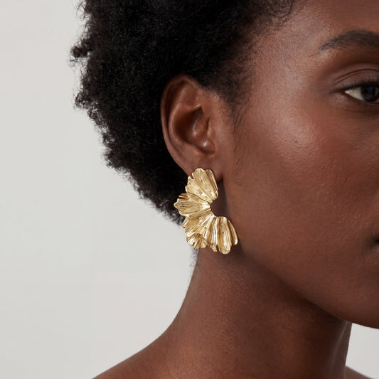 A close-up side profile of a woman with a deep skin tone wearing a large, textured gold earring shaped like a ruffled petal or ginkgo leaf with a matte metallic finish.
