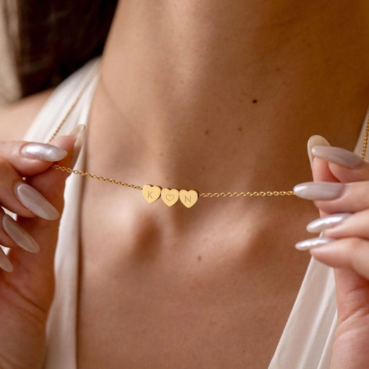 Woman holding a gold-tone triple heart initial necklace near her collarbone.