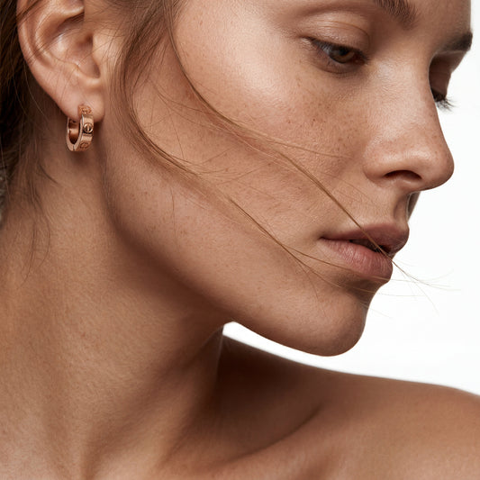 A captivating close-up portrait of a woman with natural freckles, wearing a polished rose gold hoop earring with a distinct screw motif. The model is in profile with a few hair strands blowing across her face against a pure white background.