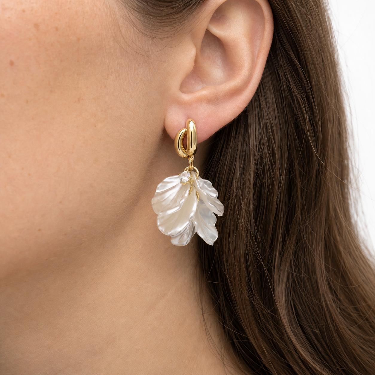 Close-up of an ear wearing a double-loop gold floral dangle earring with a clear accent stone and layered pearlescent petals.
