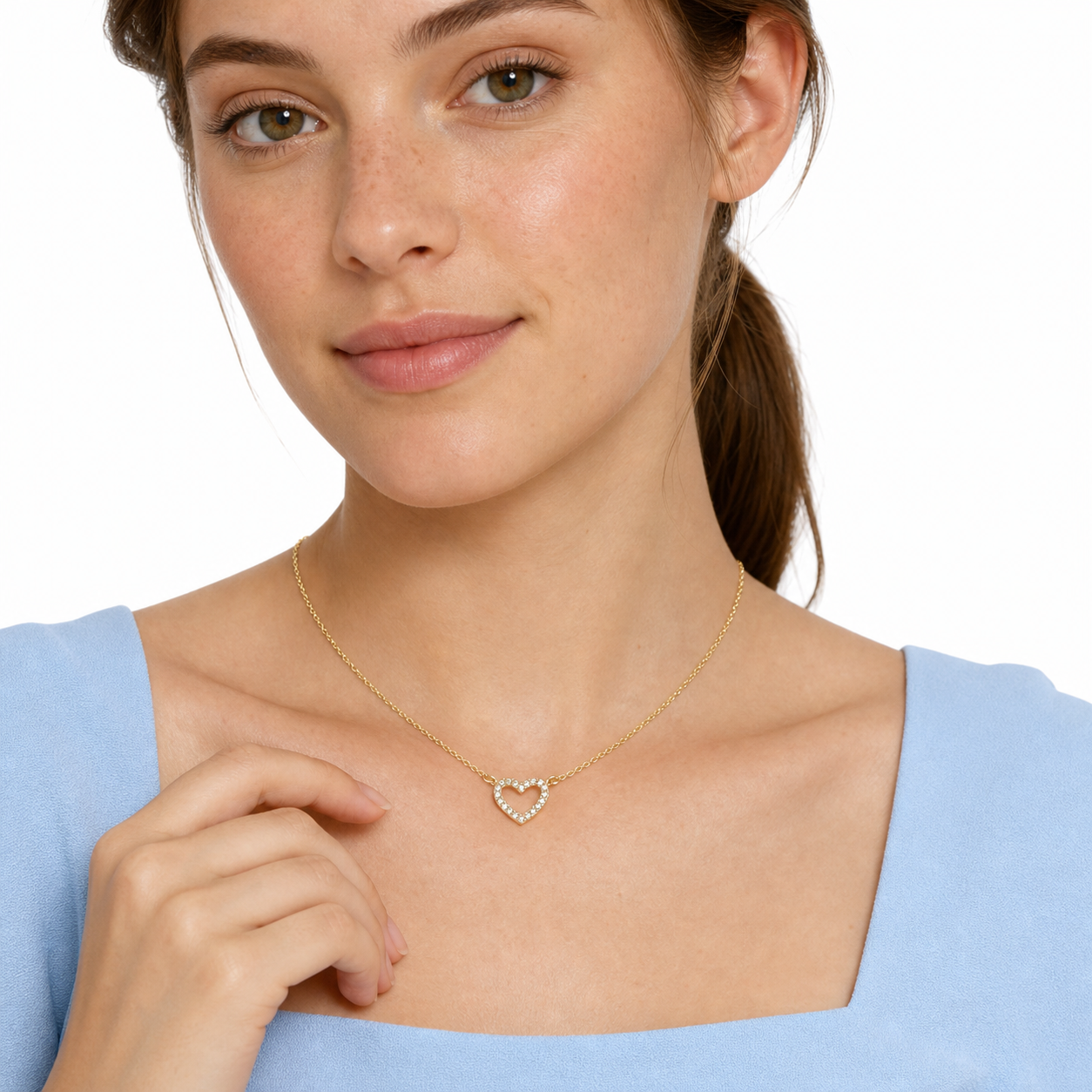 Young female model resting her hand below a gold-tone pavé open heart necklace in a white studio.