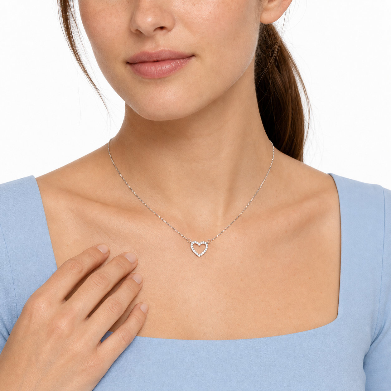 Young female model resting her hand below a silver-tone pavé open heart necklace in a white studio.
