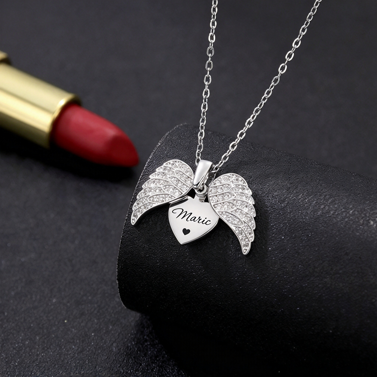 Silver-tone winged heart necklace arranged on a dark surface beside a red cosmetic item.