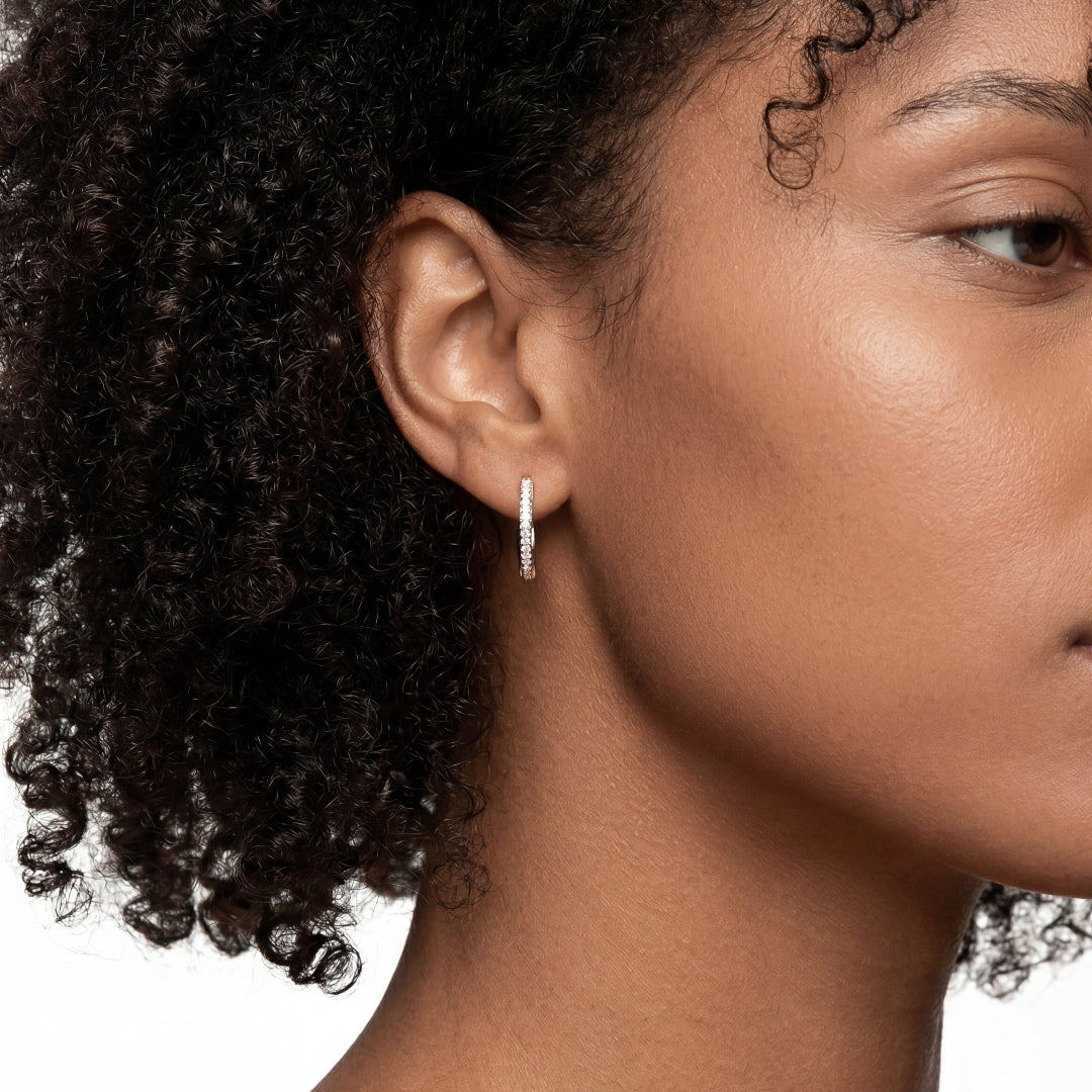 Close-up profile of a woman with dark skin and a natural curly afro, showcasing a petite, polished silver or white gold hoop earring intricately set with tiny, sparkling diamonds. High-key jewelry portrait with a pure white background.