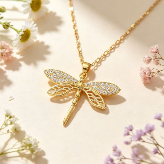 Gold-tone dragonfly pendant necklace displayed among flowers on a softly lit surface.