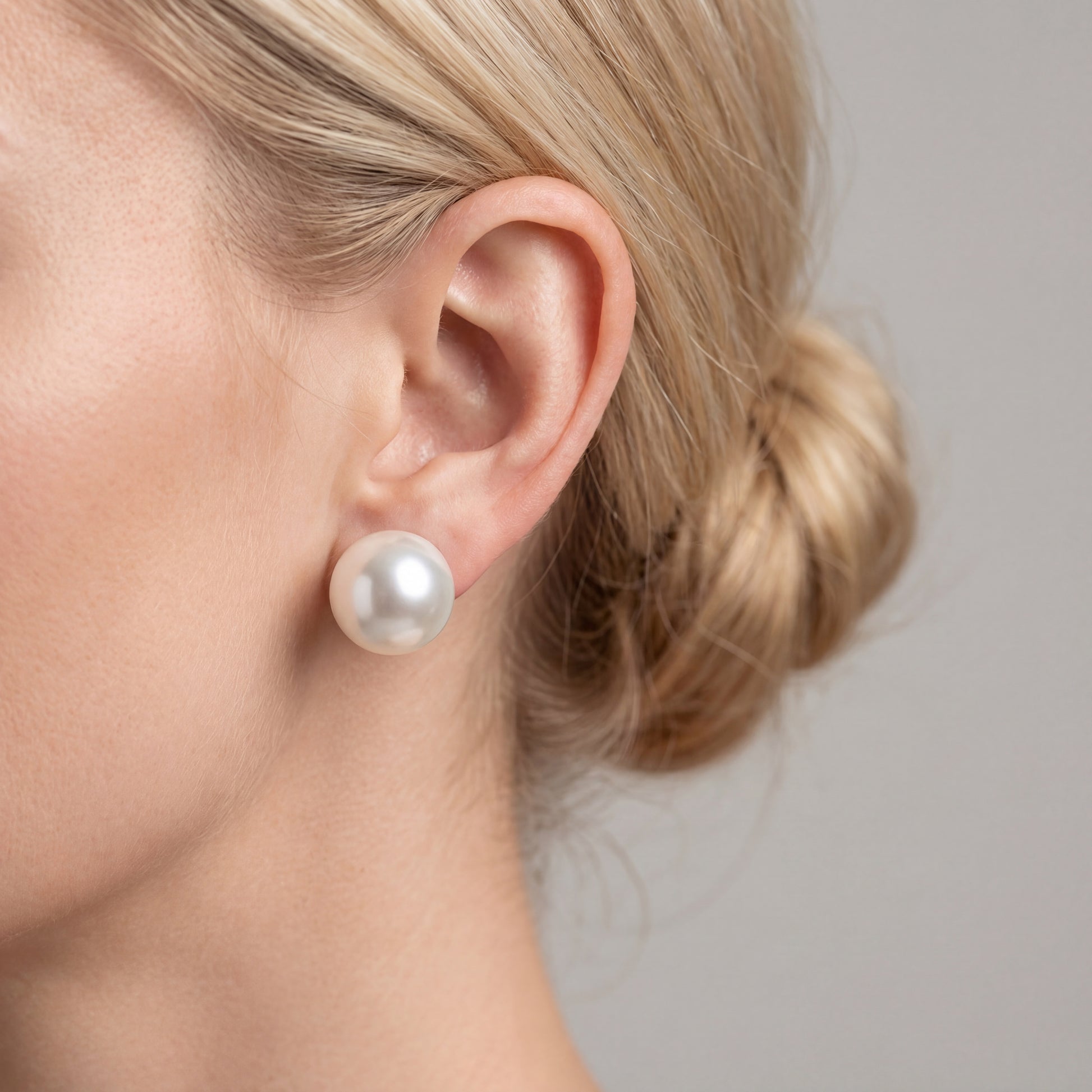 A side profile view of a fair-skinned woman with blonde hair in a chignon, wearing a single, large, high-luster white pearl stud earring against a minimalist light grey background.