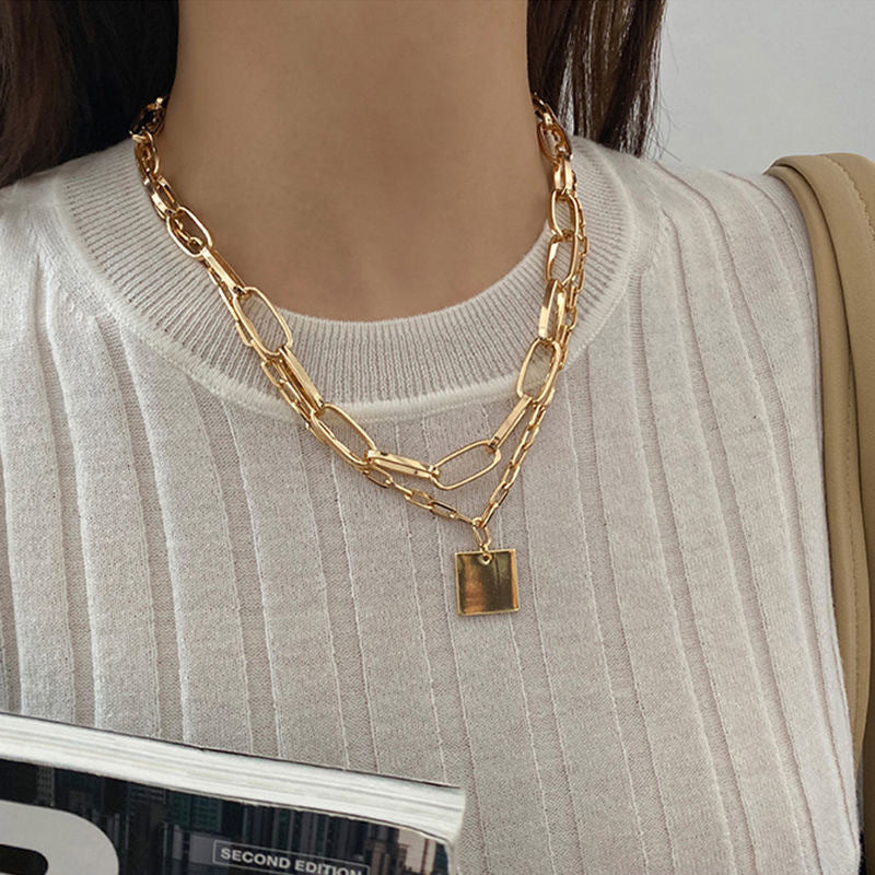 Gold-tone layered chain necklace with a square pendant worn over a light ribbed top.