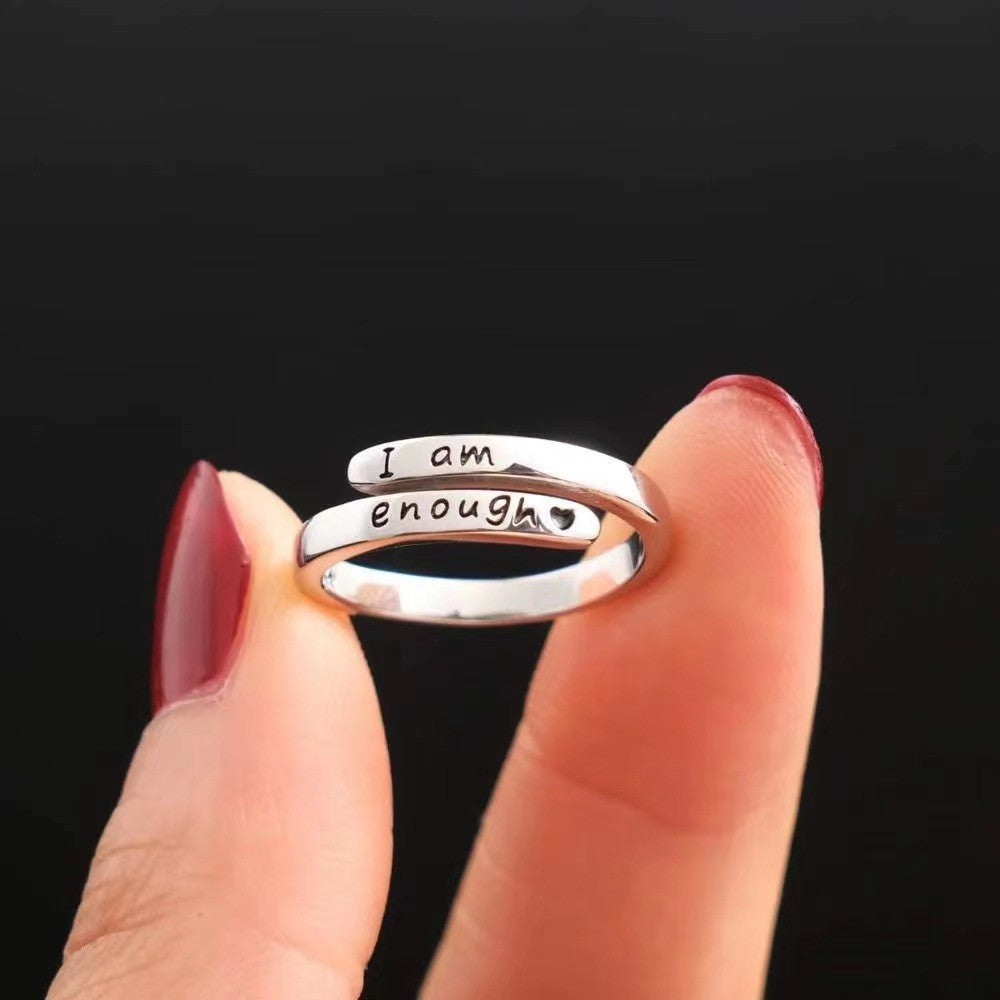 Open cuff ring displaying “I am enough” lettering between two fingertips against a dark background