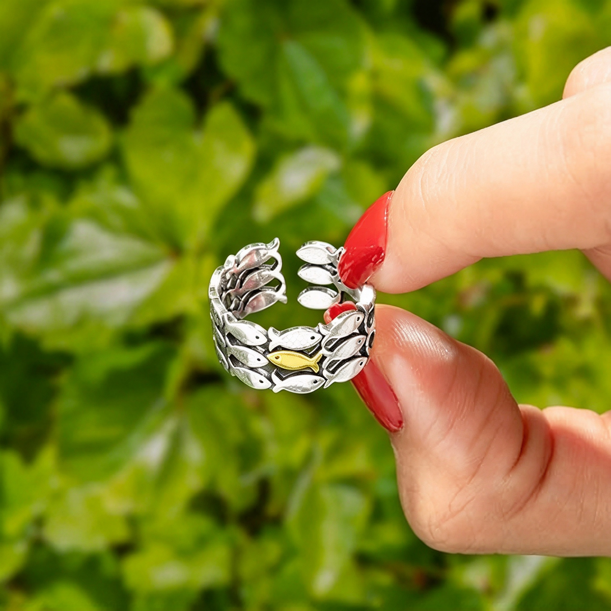 Fish school ring held between fingers against a green leafy background, highlighting the small gold-tone fish detail