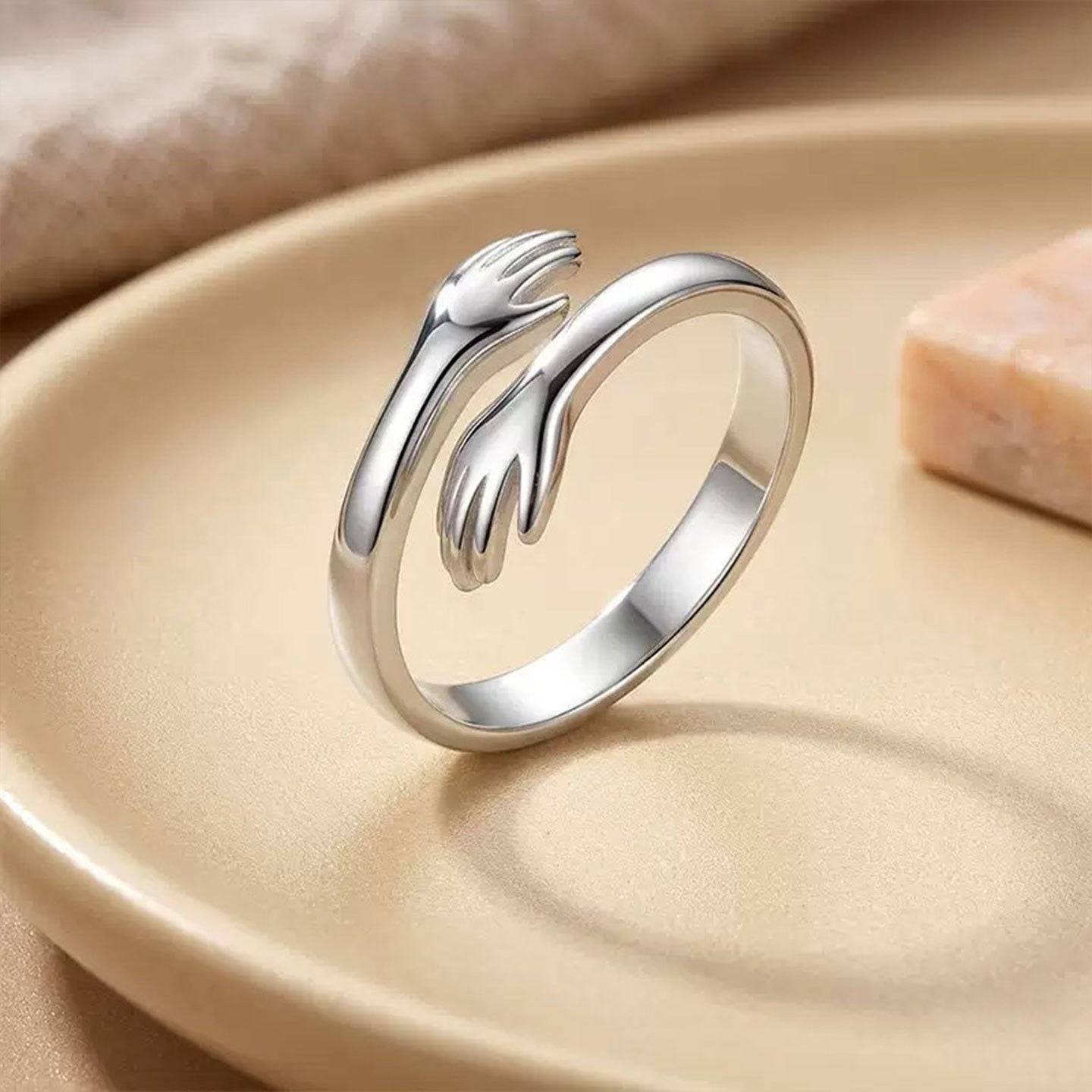 Silver-tone hugging hands ring standing upright on a warm neutral display