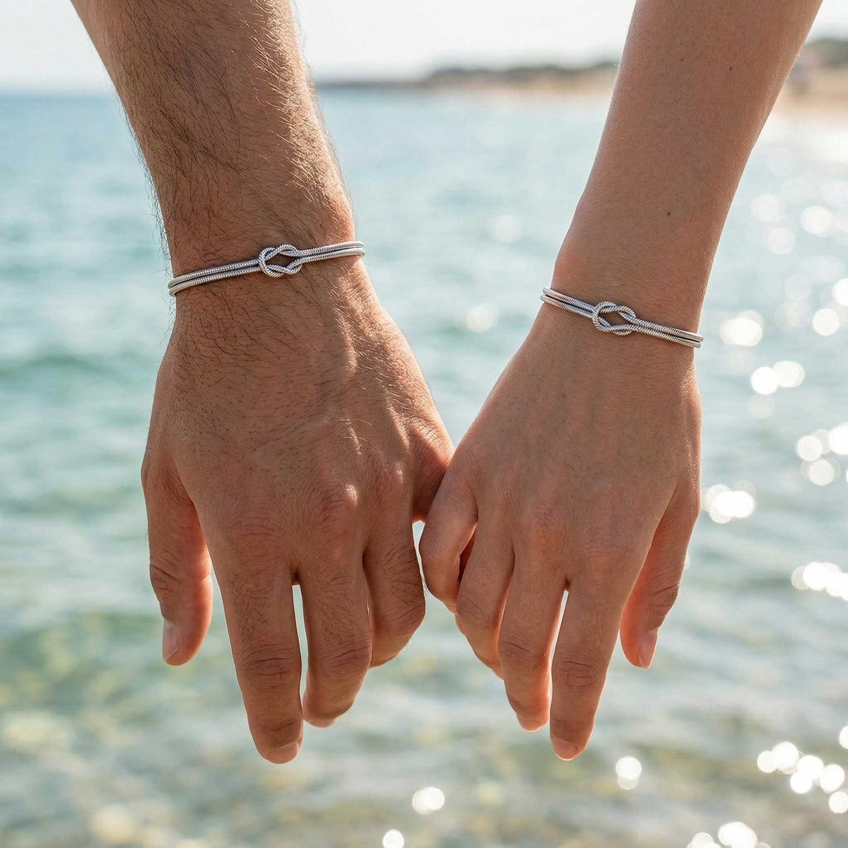Couple wearing matching knot bracelets by the sea in natural sunlight.