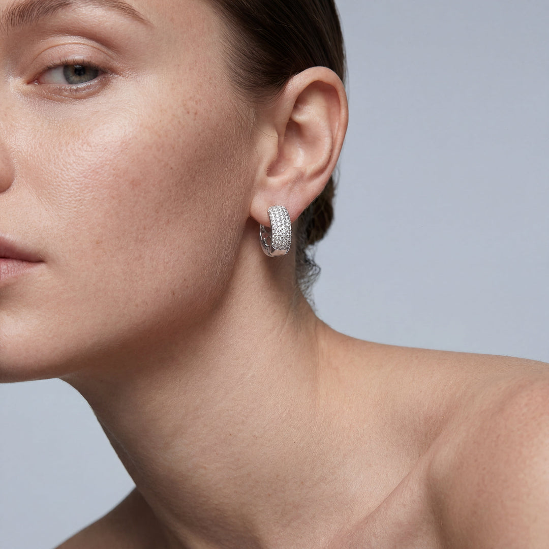 Close-up side profile of a woman wearing a wide sterling silver huggie hoop earring encrusted with sparkling pave-set diamonds. Professional jewelry photography against a neutral gray background.