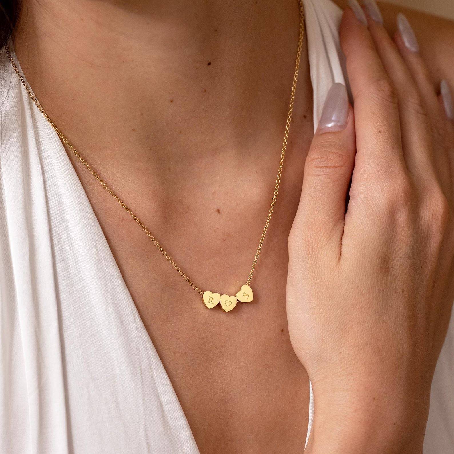 Woman wearing a gold-tone triple heart necklace with engraved initials and a white V-neck top.
