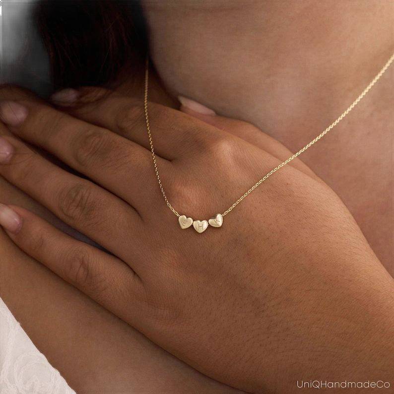 Close-up of a gold-tone three-heart initial necklace worn with a hand over the chest.