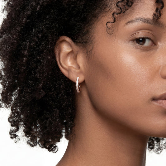 Side-profile shot of a black woman with beautiful, voluminous natural curly hair, wearing a small and elegant rose gold hoop earring adorned with a delicate row of pave-set diamonds. Professional product and model photography on a clean white backdrop.