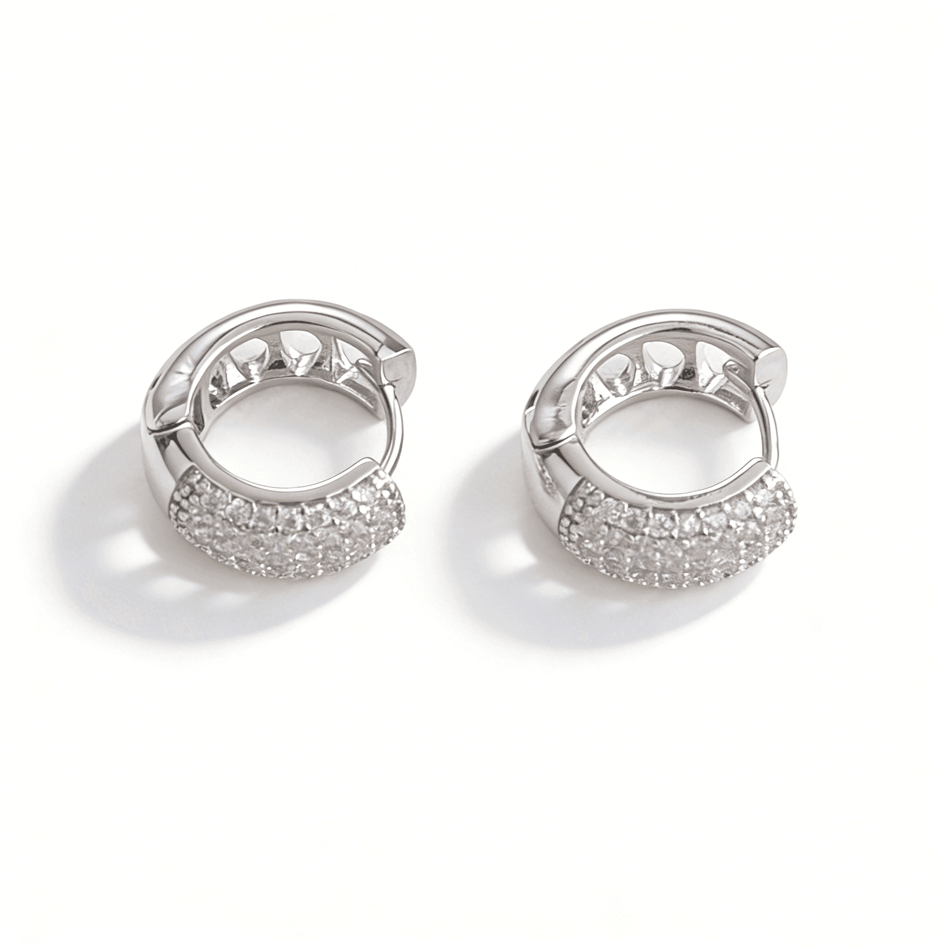 An angled overhead view of silver geometric huggie earrings, showing both the paved crystal front and the structured hollowed-out side design.
