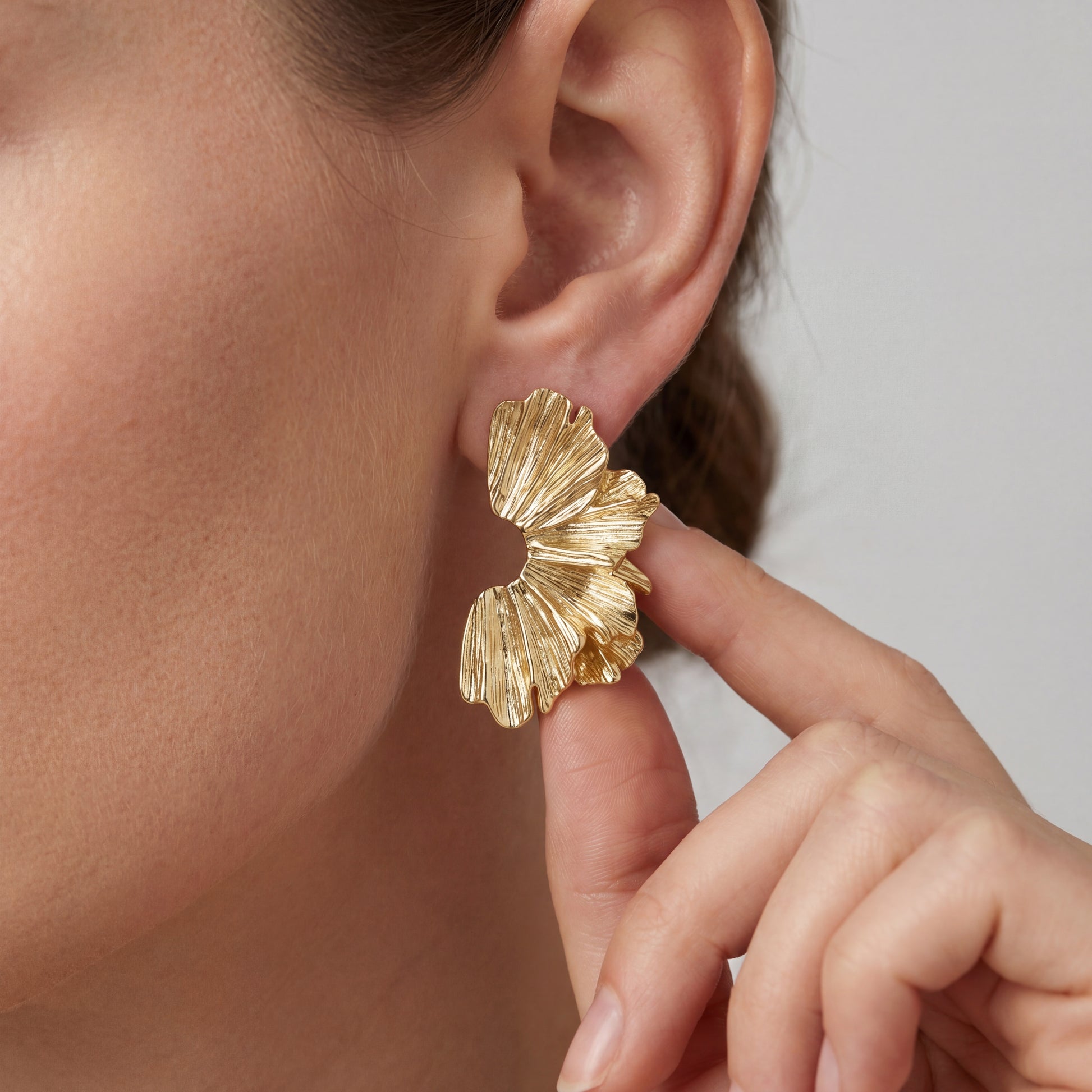 Close-up of a fair-skinned woman touching a sculptural gold fan-shaped earring, highlighting the intricate vertical ridges and organic curved edges of the jewelry.