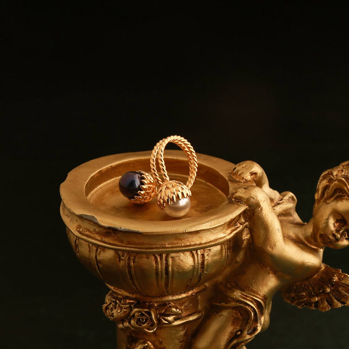 Aesthetic display of Ever P. gold pinecone pearl ring placed inside a miniature vintage golden cherub pedestal against a dark background.